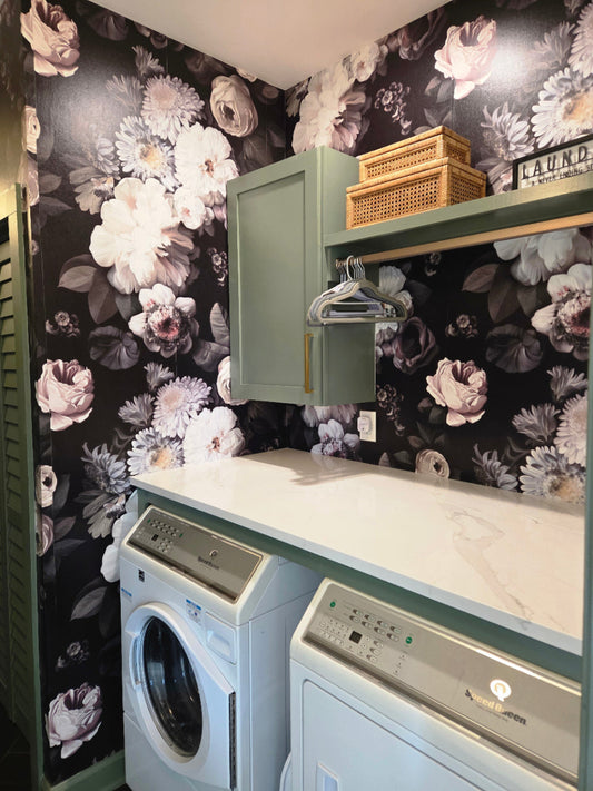 A styled laundry room corner featuring a sage green cabinet, a white marble-look countertop, a light blue washing machine, and a white dryer. The walls are covered in dramatic Dark Floral Wallpaper by Ellie Cashman with large cream and gray roses.