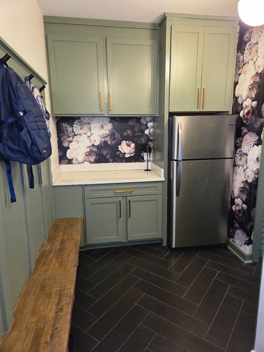 A laundry room with sage green cabinets, a stainless steel refrigerator, dark herringbone tile flooring, a wooden bench, and Dark Floral Wallpaper by Ellie Cashman.