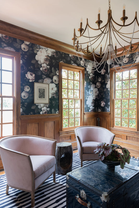 Dark Floral wallpaper on all walls above oak wainscoting in living room with pink chairs, brass chandelier, and striped rug