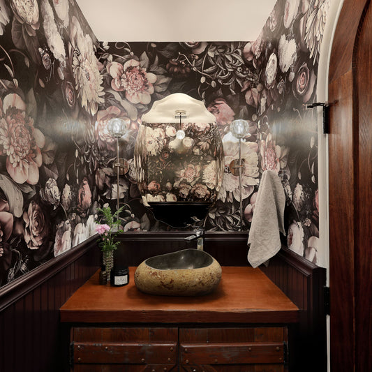 A bathroom corner featuring Ellie Cashman's dramatic dark floral wallpaper in black, beige, and dusty rose tones, with a dark wood vanity, a stone vessel sink, and a wooden door on the right.
